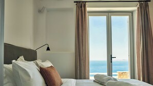 A hotel room with a bed, a nightstand, a lamp, and a view of the sea through a large window. - Hotiday Mykonos Fanari (Mykonos)