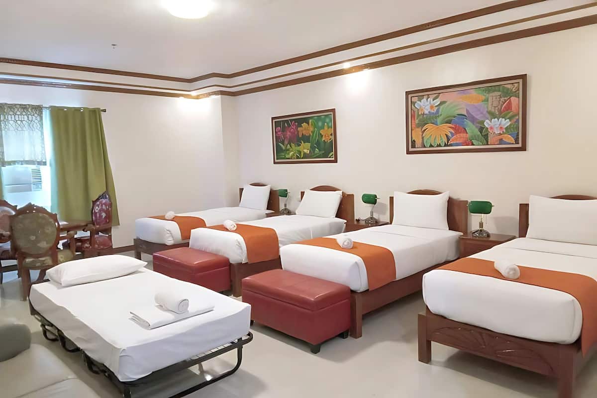 A hotel room with four beds, each with white and orange bedding, a small table with a lamp, and artwork on the walls.