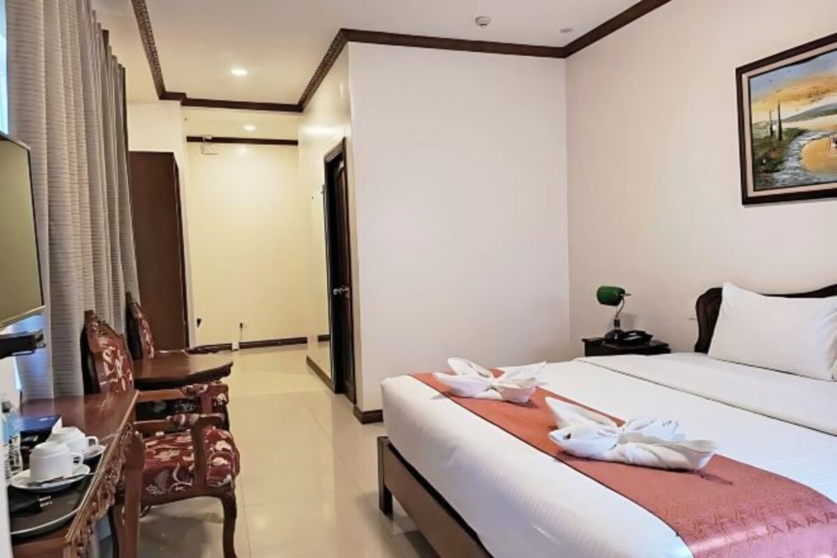 A hotel room with a bed, a desk with a chair, a dining table, and a painting on the wall.