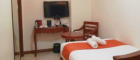 Habitación de hotel con una cama, un escritorio con televisor, una silla y un cesto de basura pequeño.