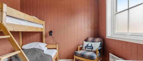Une petite pièce agréable et chaleureuse, avec un lit superposé, une chaise dotée d’un coussin à carreaux, une lampe fixée au mur et une fenêtre.