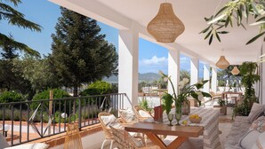 A sunlit outdoor seating area with wicker furniture, a wooden table, and a view of the surrounding landscape.