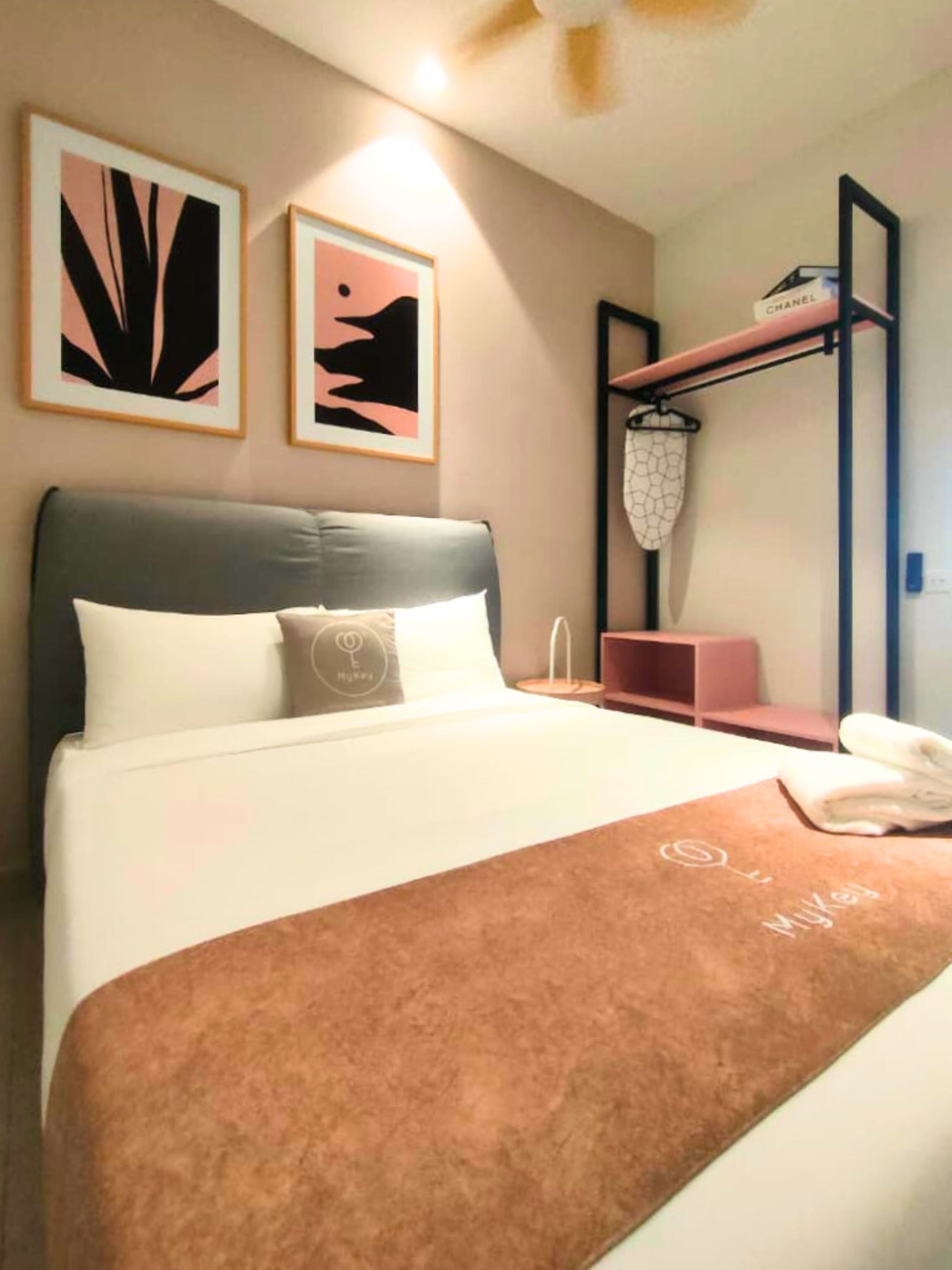 Quarto de hotel com cama, mesa de cabeceira, prateleira com cesto pendurado e duas pinturas abstratas emolduradas na parede.