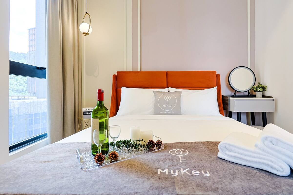 A hotel room with a bed, a bedside table with a mirror, a bottle of wine, glasses, and a tray with pine cones.