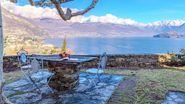 A stone table with chairs, a book, and a bowl of fruit in a scenic outdoor setting with a view of mountains and a lake. - Stunning private villa for 4 guests with WIFI, TV and patio (Calozzo)