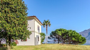 A white building with a stone wall in front, a grassy area, and a mountain in the background. - Stunning private villa for 4 guests with WIFI, TV and patio (Calozzo)