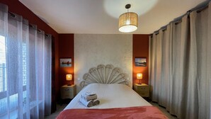Une chambre d’hôtel avec un grand lit, deux tables de chevet, une tête de lit décorative et une vue sur la mer par la fenêtre.