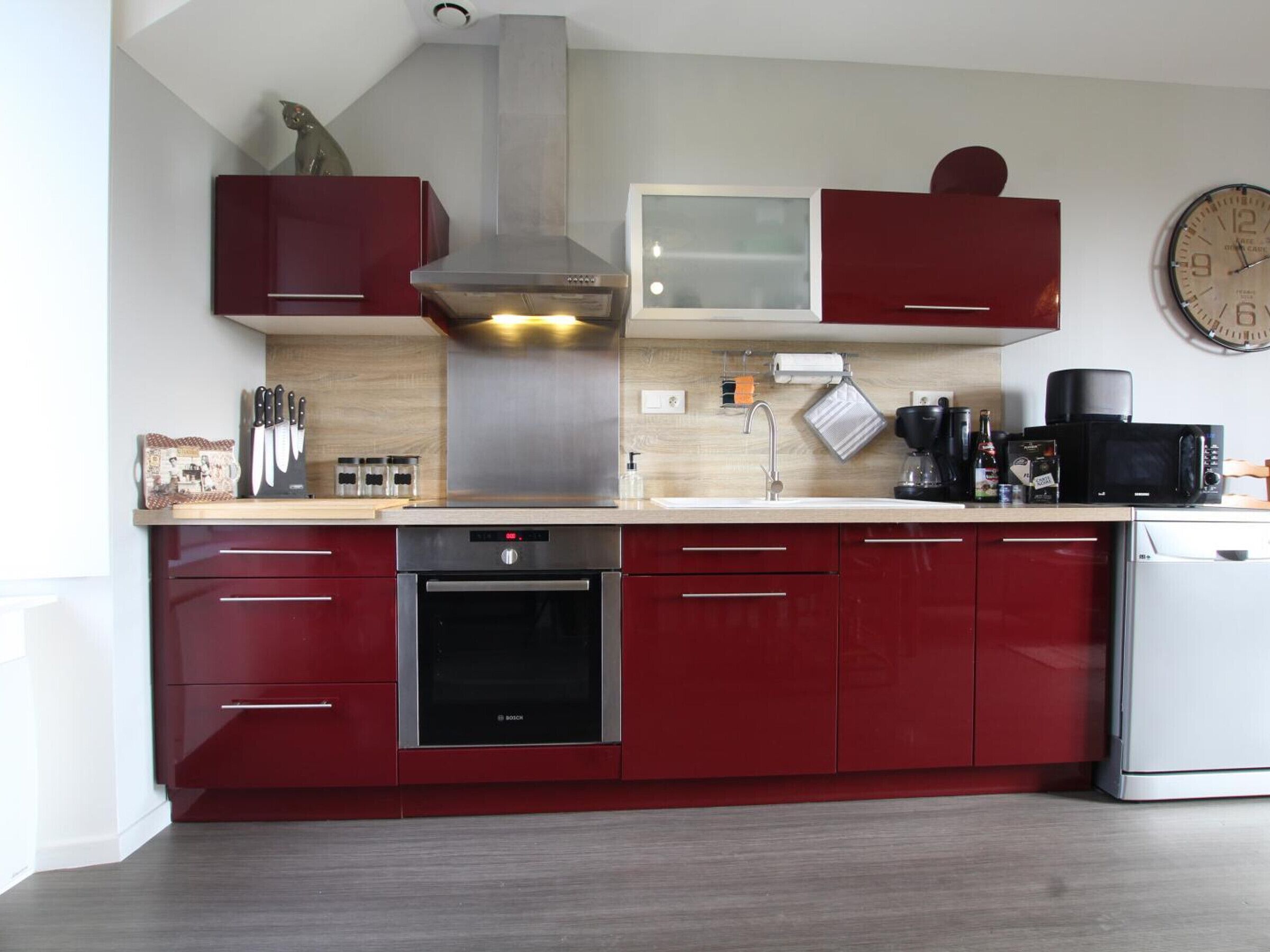 A modern kitchen with red cabinets, stainless steel appliances, and a clock on the wall.