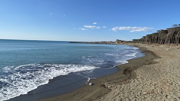 Une plage avec un ciel dégagé, une mer calme et un promontoire rocheux où se trouvent des bâtiments au loin.