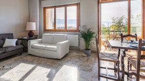 Un salon avec un canapé blanc, une petite table avec une lampe et une vue sur l’extérieur grâce à de grandes fenêtres.