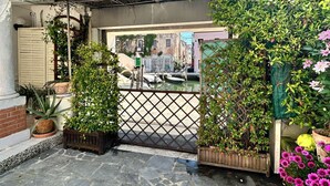 Une entrée fermée par un portail en métal, agrémentée de plantes en pot et d’une allée pavée menant à un bâtiment doté d’un balcon.