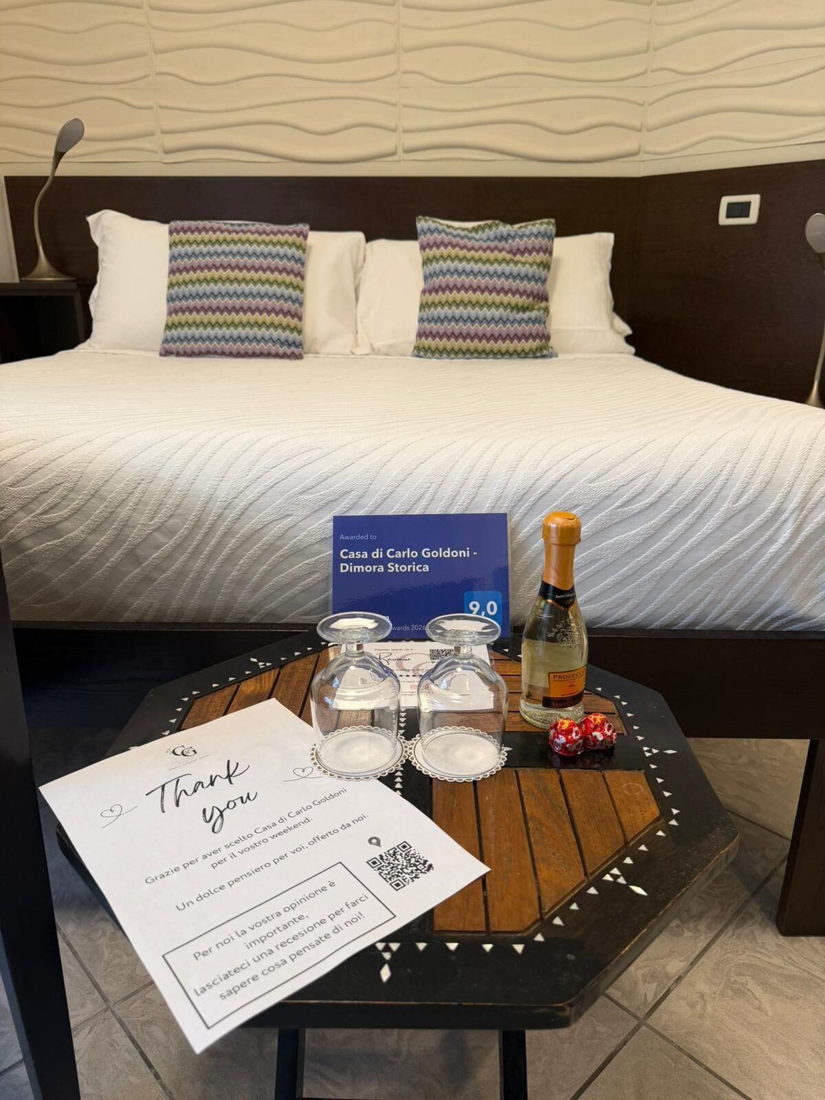 Una cama bien hecha con ropa blanca y almohadas coloridas, una mesita de noche con una nota de agradecimiento, una botella de Prosecco y una pequeña caja de regalo.