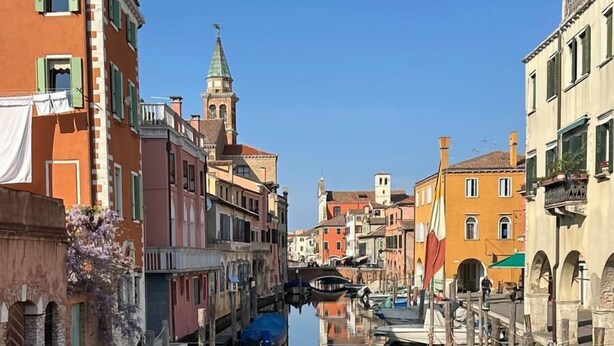 Un canal bordé de bâtiments des deux côtés, des bateaux amarrés le long de l’eau et, au loin, le clocher d’une église.
