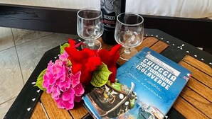 Un lit bien fait, avec du linge de lit blanc, une bouteille de vin, deux verres et un livre posés sur une table en bois.