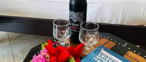 A neatly made bed with white linens, a bottle of wine, two glasses, and a book on a wooden table.