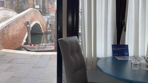 Une chambre avec vue sur un canal et des bâtiments historiques, une chaise, une table avec un pied bleu et une pancarte indiquant le nom de la chambre et son prix.