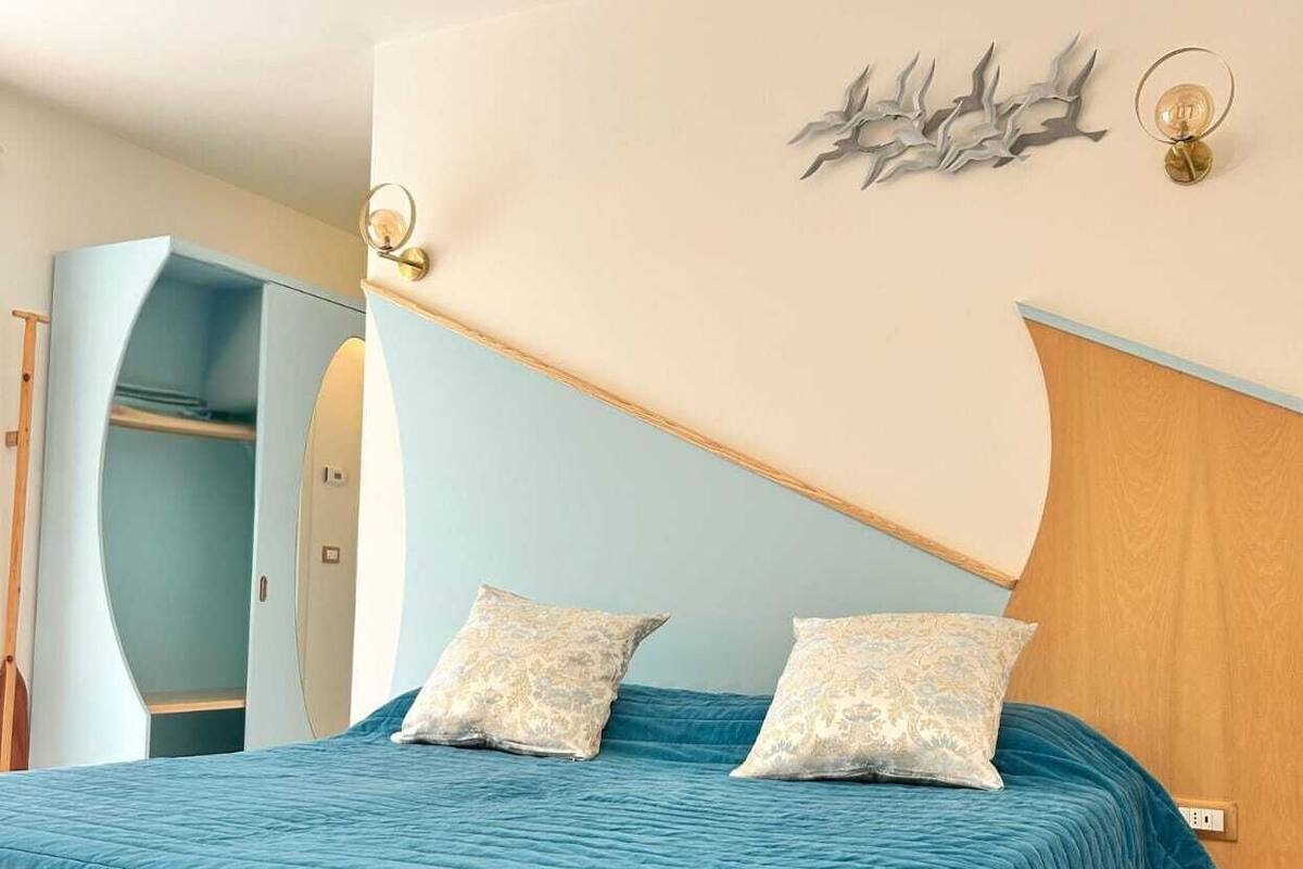 Een slaapkamer met een bed, twee kussens, een nachtkastje, een wandlamp en een deur naar een andere kamer.