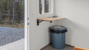 Esquina de una habitación con un estante de madera, un cubo de basura y una puerta que da a un área de grava.