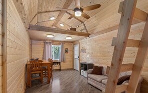 A cozy cabin interior with a dining area, a sofa, a ceiling fan, and a fireplace. - Red Cabin at Fish Camp – Cozy Dry Cabin with Loft | Rustic Escape (Sterling)