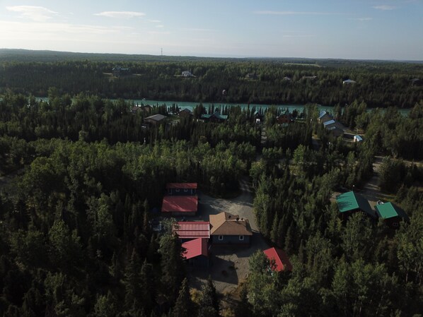 Luftaufnahme eines bewaldeten Gebiets mit mehreren Gebäuden, darunter Blockhäuser und Häusern, umgeben von dichtem Wald und einem See.