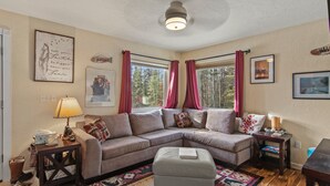 A cozy living room with a sectional sofa, a small table, a lamp, and a window with curtains. - King Cabin -Full Kitchen + Laundry | Kenai Retreat (Sterling)