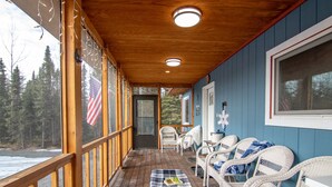 A covered porch with wooden railings, wicker furniture, and a view of a forested area.