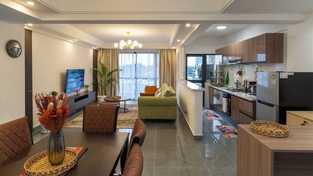 A modern living room with a sofa, dining table, and a TV. There is a kitchen area with cabinets and a refrigerator. - Skyline symmetry (Nairobi)