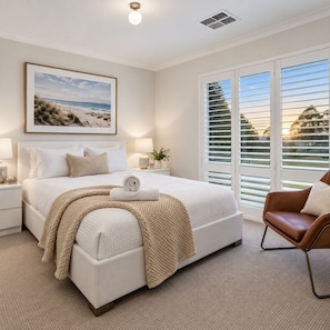 A bedroom with a bed, bedside tables, a nightstand, a chair, a window with blinds, and a picture on the wall.