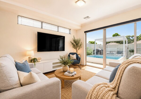 A modern living room with a flat-screen TV, comfortable seating, a coffee table, and a view of a backyard with a pool.