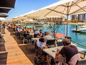 A waterfront dining area with tables and chairs, a wooden deck, and a view of the marina with boats. - Cozy Coastal Retreat | Marina, Beach & Plunge Pool (Mandurah)