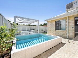 A swimming pool with a pergola, a brick house, and a fenced backyard.