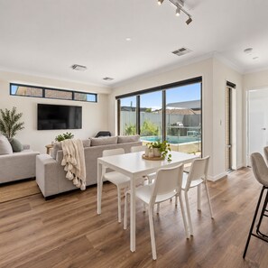 A modern living room with a sofa, a dining table, and chairs. There is a TV mounted on the wall and a large window providing a view of the outdoors.