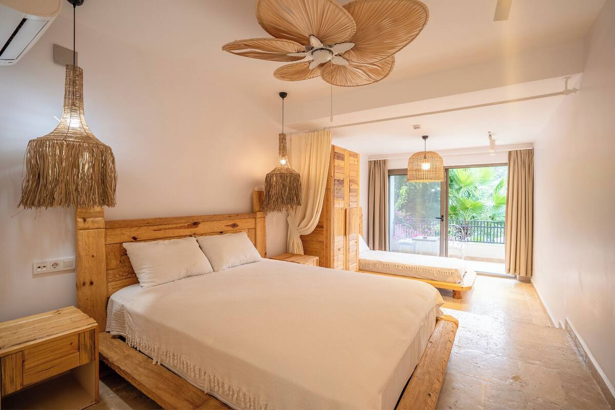 A bedroom with a wooden headboard, a large bed, two hanging wicker lamps, a ceiling fan, and a window with a view of greenery.