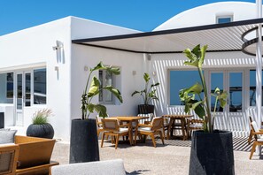 A modern outdoor seating area with a white building, potted plants, and wooden furniture. - Oikos Santorini Living Concept (Santorini)