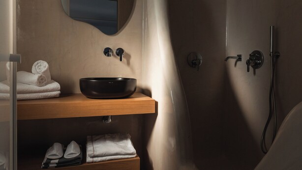 A bathroom with a wooden vanity, a round mirror, a black sink, and a walk-in shower. - Oikos Santorini Living Concept (Santorini)