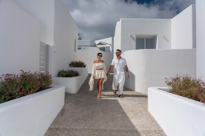 A couple walking on a white-walled pathway with red flowers and a clear sky. - Oikos Santorini Living Concept (Santorini)