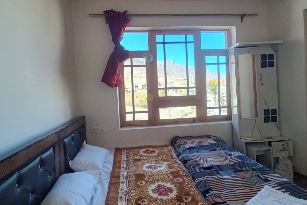 A bedroom with a wooden ceiling, a large window with a view of mountains, a bed with a patterned bedspread, and a desk with a computer.