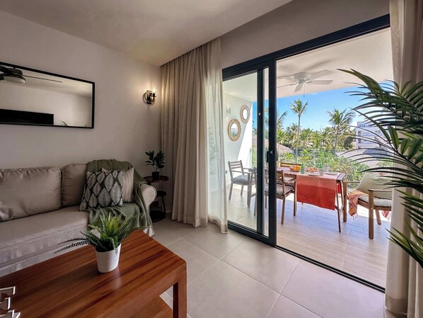 Una sala de estar moderna con un sofá, una mesa de centro y un comedor con una mesa y sillas. Hay una puerta corrediza de vidrio que da a un patio al aire libre con una mesa dispuesta para comer y vistas a palmeras y un edificio.