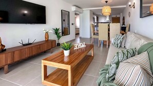 A modern living room with a wooden coffee table, a sofa with striped pillows, a TV mounted on the wall, and a dining area in the background.