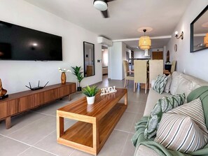 A modern living room with a wooden coffee table, a sofa with striped pillows, a TV mounted on the wall, and a dining area in the background. - Beautiful new condo with Picuzzi (Punta Cana)