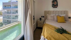 A hotel room with a bed, a nightstand, a small potted plant, and a view of a swimming pool and buildings.