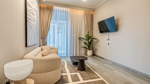 Een moderne woonkamer met een beige bank, een ronde salontafel, een vloerlamp, een aan de muur bevestigde televisie en een groot raam met dunne gordijnen.