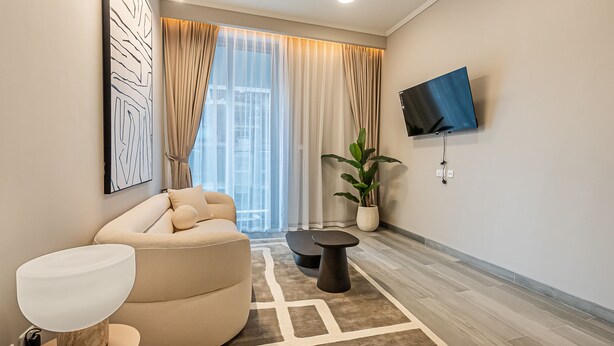 Un salon moderne avec un canapé beige, une table basse ronde, un lampadaire, un téléviseur fixé au mur et une grande fenêtre ornée de rideaux légers.