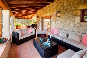 A covered outdoor seating area with wicker furniture, stone walls, and a view of the garden.