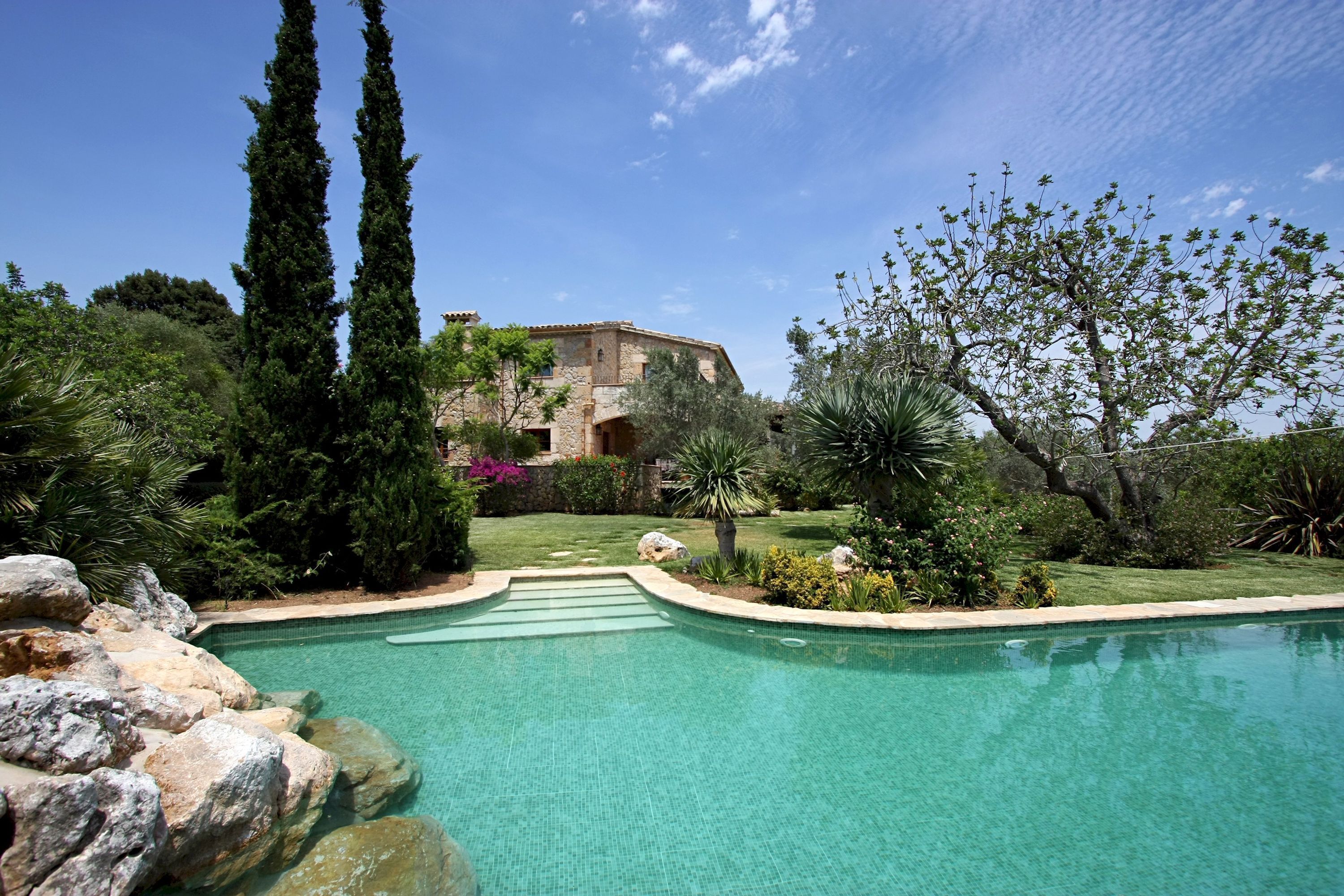 A swimming pool with a stone border, surrounded by rocks and greenery, with a building in the background.