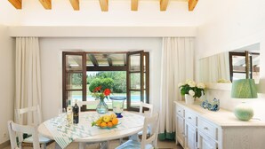 Un coin repas avec une table ronde, des chaises, un vase de fleurs, un saladier rempli de fruits et une bouteille de vin.