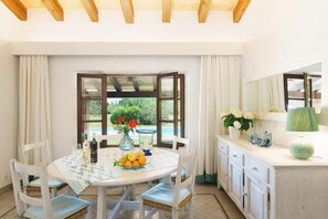 A dining area with a round table, chairs, a vase with flowers, a bowl of fruit, and a bottle of wine. - Villa Reco Verd in Pollen a (Pollenca)
