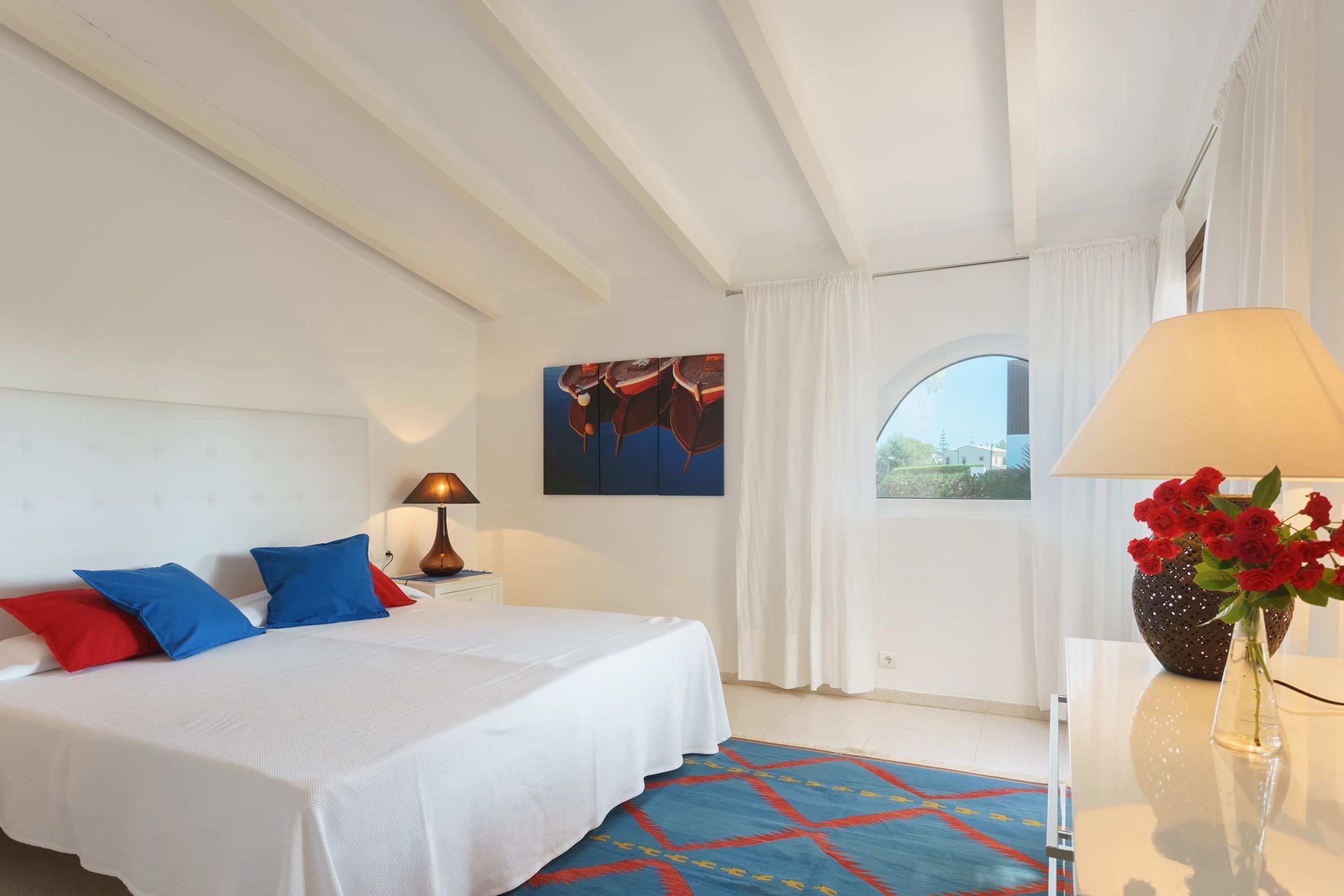 A hotel room with a bed, a nightstand, a lamp, a vase with flowers, a painting on the wall, and a window with curtains.