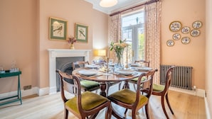 Un comedor con una mesa redonda dispuesta para cuatro, rodeada de sillas de madera con cojines verdes. Se observa una chimenea con repisa blanca al fondo, y cuadros enmarcados en la pared.