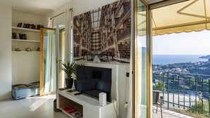 A modern living room with a large window, a flat-screen TV, a bookshelf, and a view of a coastal landscape. - Elegant sea View Apartment by Wonderful Italy (Rapallo)